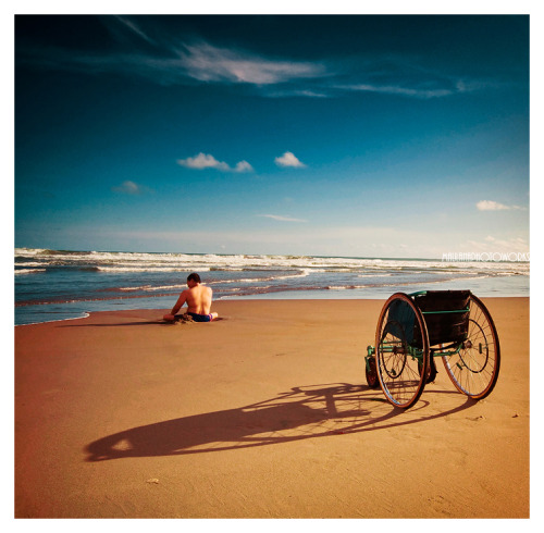 An image of a beach with blue sky, dotted with clouds. A figure sits in the sand by the waves. In the foreground, a wheelchair sits on the sand.
